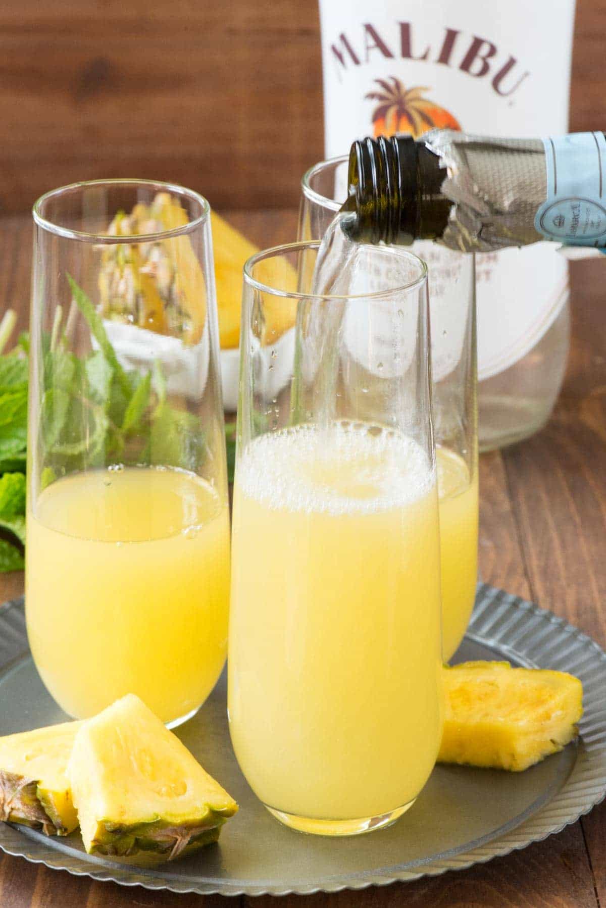 Adding Prosecco into three drinking glasses to make Hawaiian Mimosas .