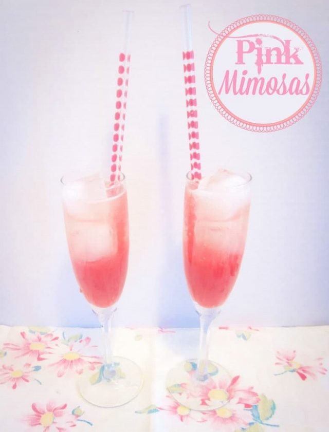 Two champagne glasses of Pink Mimosas with pink straws on a floral tablecloth. 