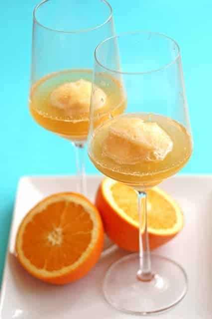 Two wine glasses of Orange Sorbet Mimosa on a white serving plate with sliced oranges. 