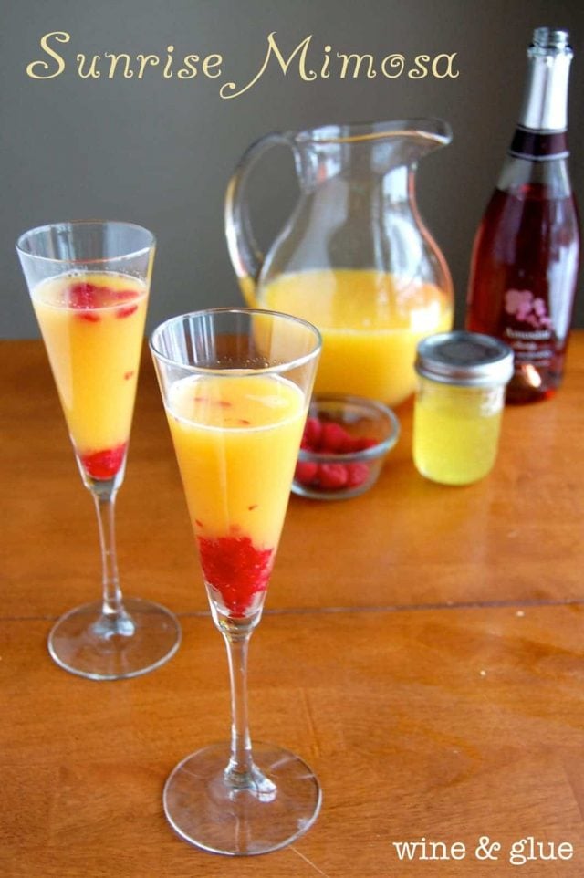 Two glasses of Sunrise Mimosas with a pitcher, champagne and fresh raspberries in the background. 