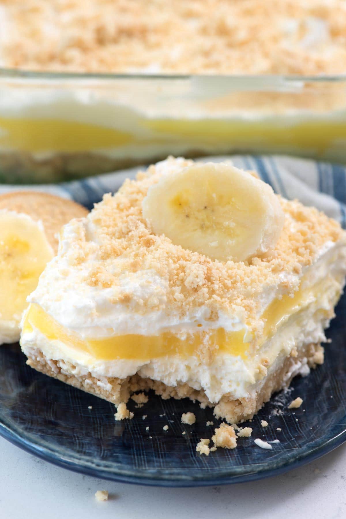 Slice of No Bake Banana Pudding Dream Dessert on a dark blue plate with the rest of the cake in a glass dish in the background