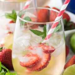 2 Clear glass half filled with Strawberry Lime Punch with ice. Strawberries are in the glass and it has some mint and a red and white stripes. Thee are lime slices and mint and strawberries next to the glasses