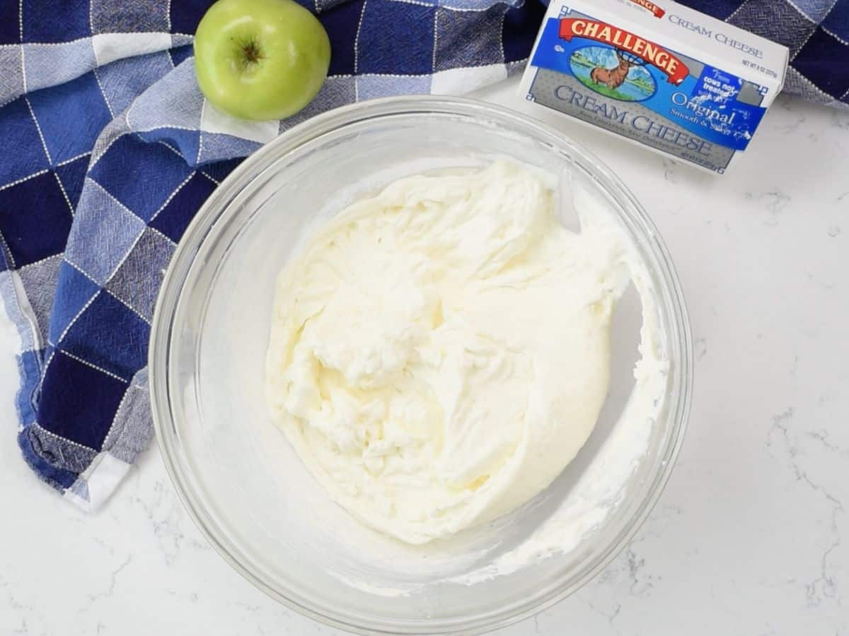 bowl of cheesecake mixture with cream cheese box and apple behind
