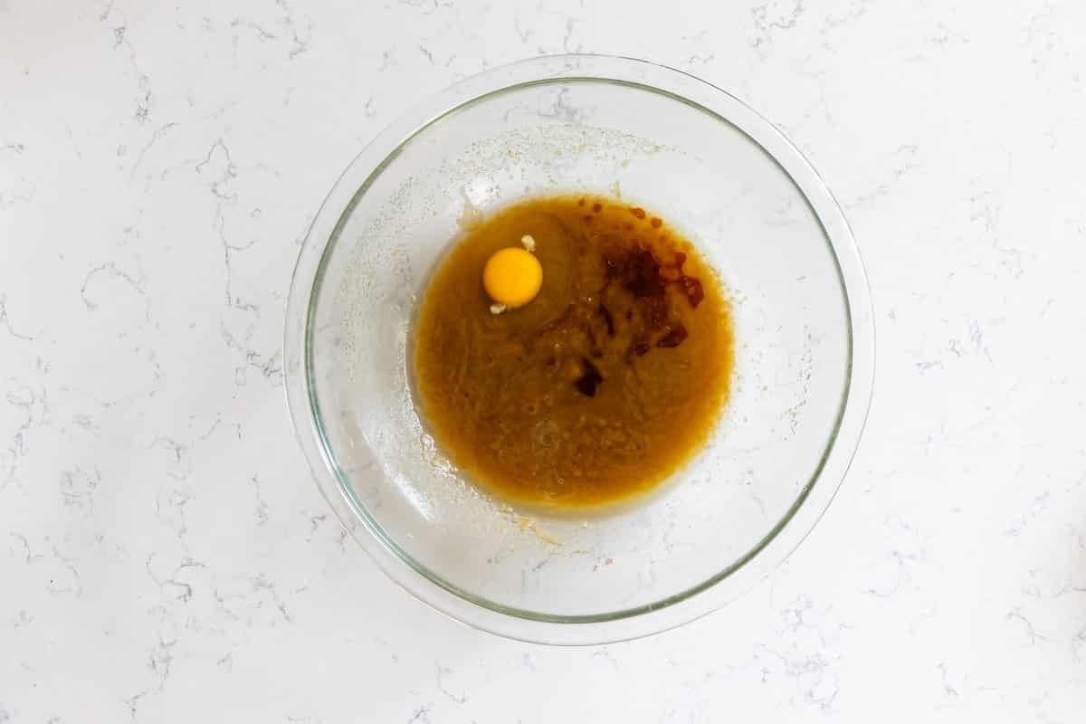 A glass mixing bowl on a white countertop contains brown liquid, an egg, and some vanilla extract—ingredients ready to be mixed for baking classic Doubletree Chocolate Chip Cookies.