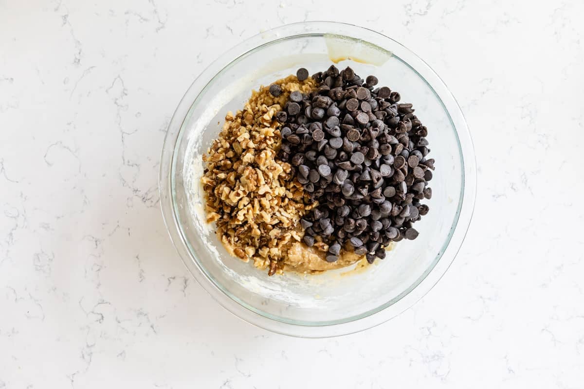 A glass mixing bowl on a white countertop holds batter topped with chopped walnuts on one side and chocolate chips on the other, ready to be mixed—perfect for homemade Doubletree Chocolate Chip Cookies.