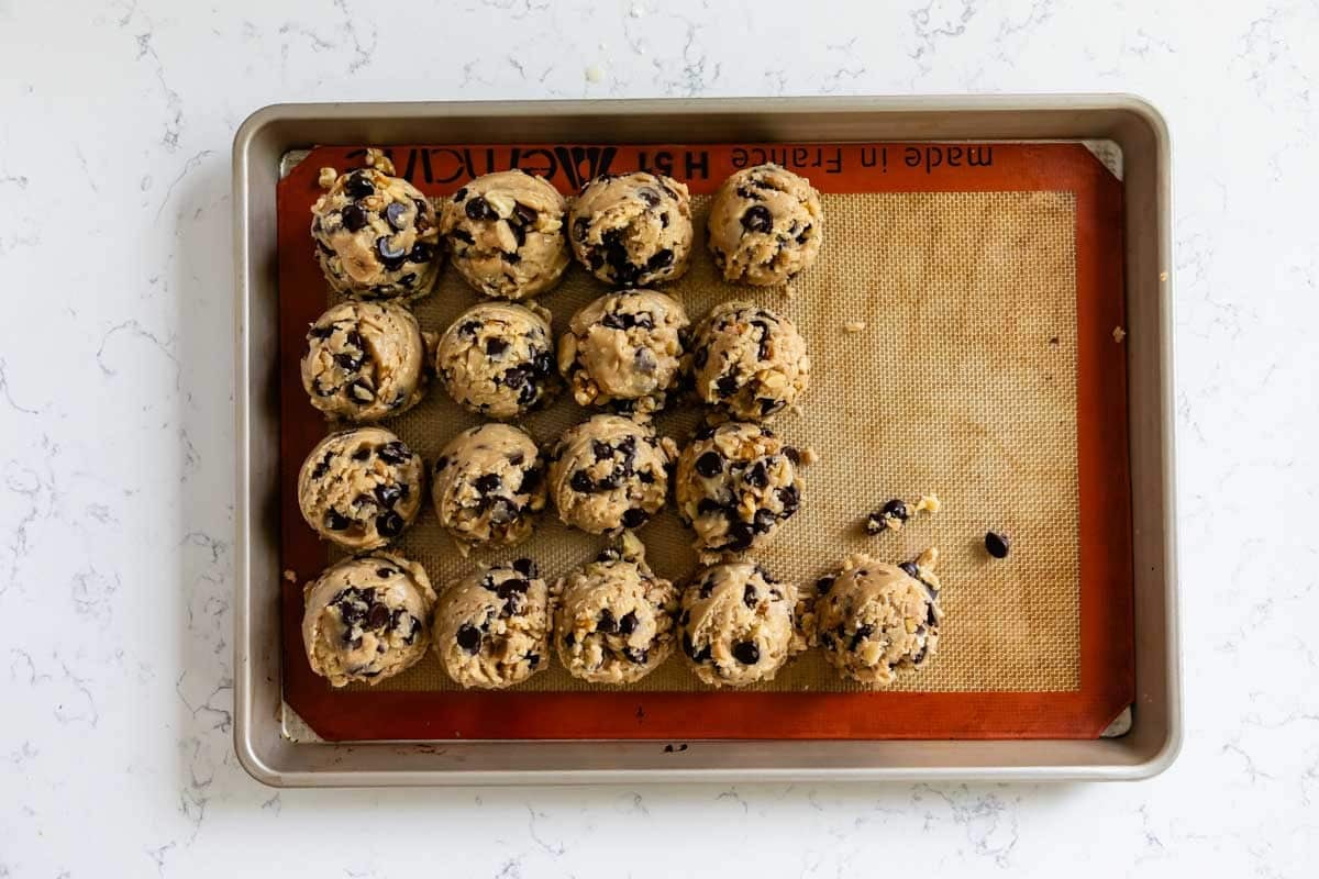 A baking sheet lined with a silicone mat holds 16 scoops of Doubletree Chocolate Chip Cookies dough arranged in rows, ready to be baked, on a white countertop.