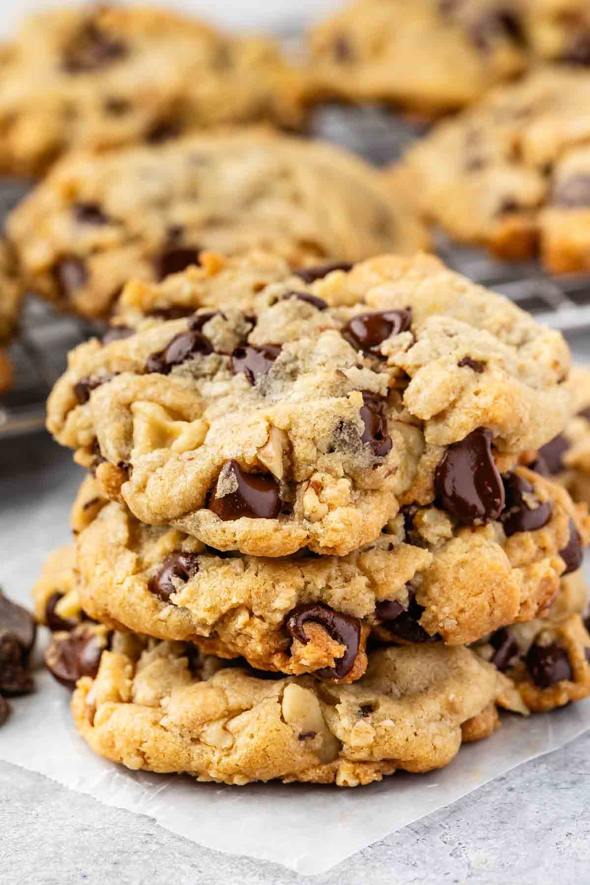 A stack of three thick, homemade Doubletree Chocolate Chip Cookies with gooey chocolate chunks rests on parchment paper, with more cookies blurred in the background.