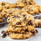 A stack of three Doubletree Chocolate Chip Cookies sits on parchment paper, with more cookies and scattered chocolate chips in the background on a wire cooling rack. The cookies have a golden-brown, chunky appearance.