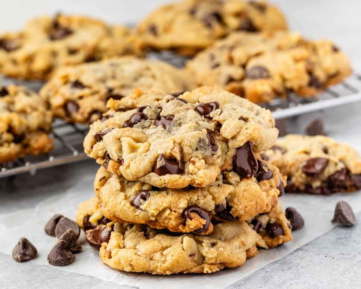 A stack of three Doubletree Chocolate Chip Cookies sits on parchment paper, with more cookies and scattered chocolate chips in the background on a wire cooling rack. The cookies have a golden-brown, chunky appearance.
