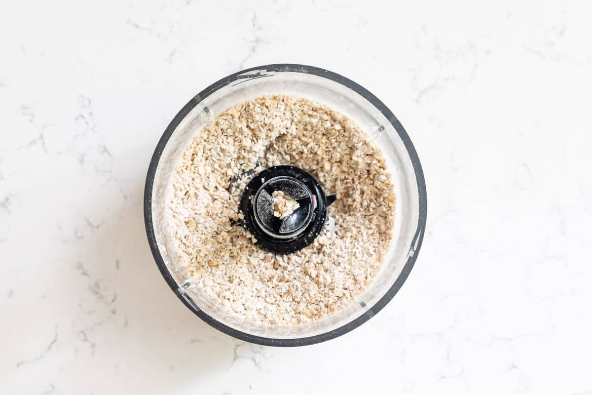 A food processor bowl filled with finely ground oats—perfect for making Doubletree Chocolate Chip Cookies—sits on a white marble countertop, viewed from above.