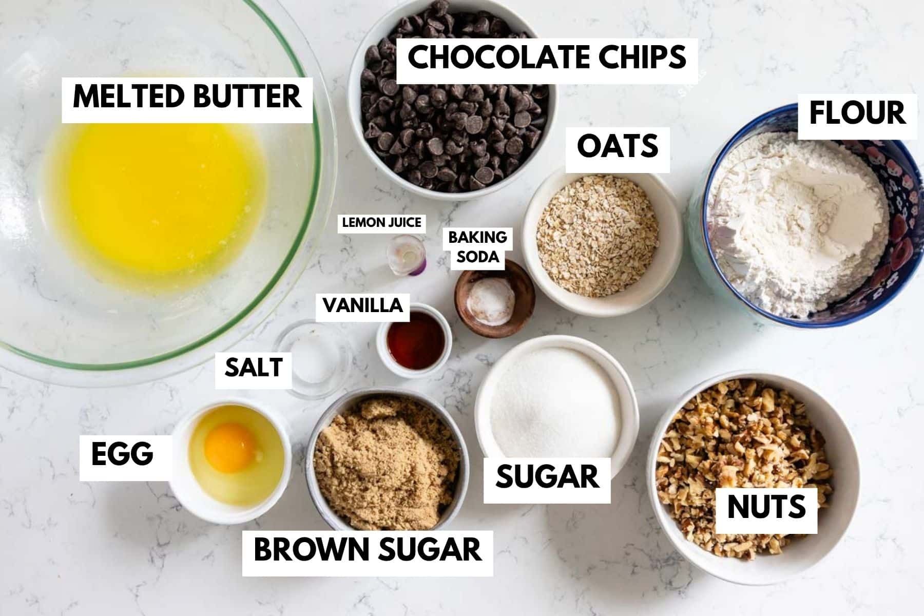 Top-down view of labeled Doubletree Chocolate Chip Cookies ingredients in bowls, including melted butter, chocolate chips, oats, flour, egg, brown sugar, sugar, nuts, salt, vanilla, lemon juice, and baking soda on a white surface.