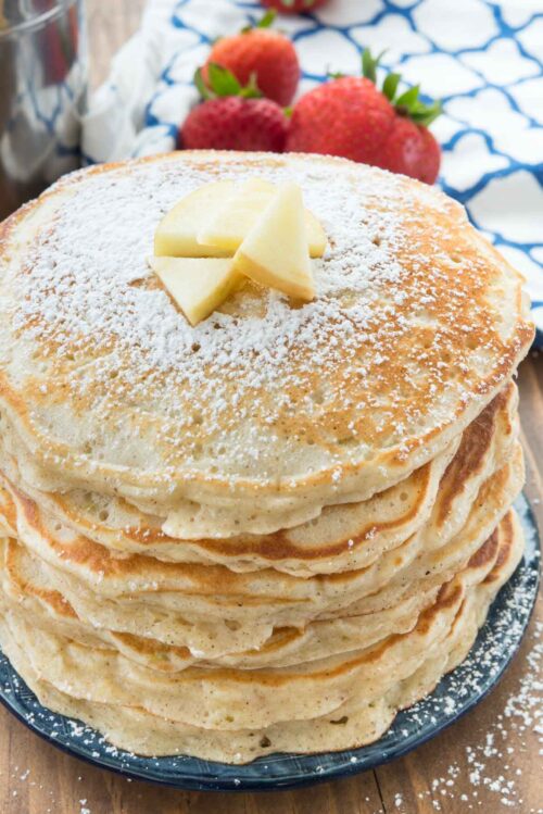 Stack of pancakes with powdered sugar and butter on blue plate with strawberries and a blue and white napkin