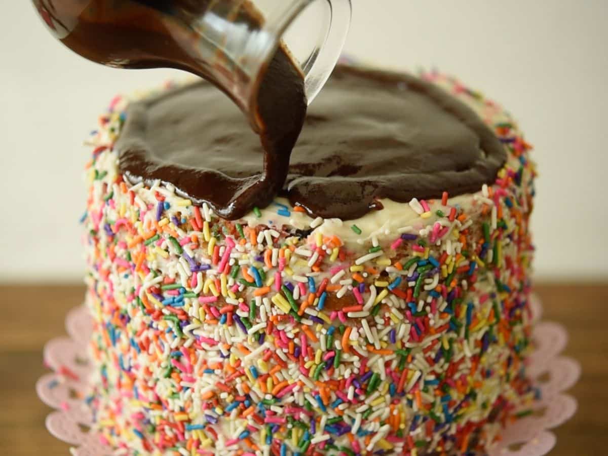 cake on cake plate with sprinkles around it and fudge topping pouring over it.