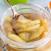 Sliced cooked apples with cinnamon in a glass jar, being scooped with a spoon. The soft, syrupy apple pie filling glistens, with visible flecks of spice throughout.