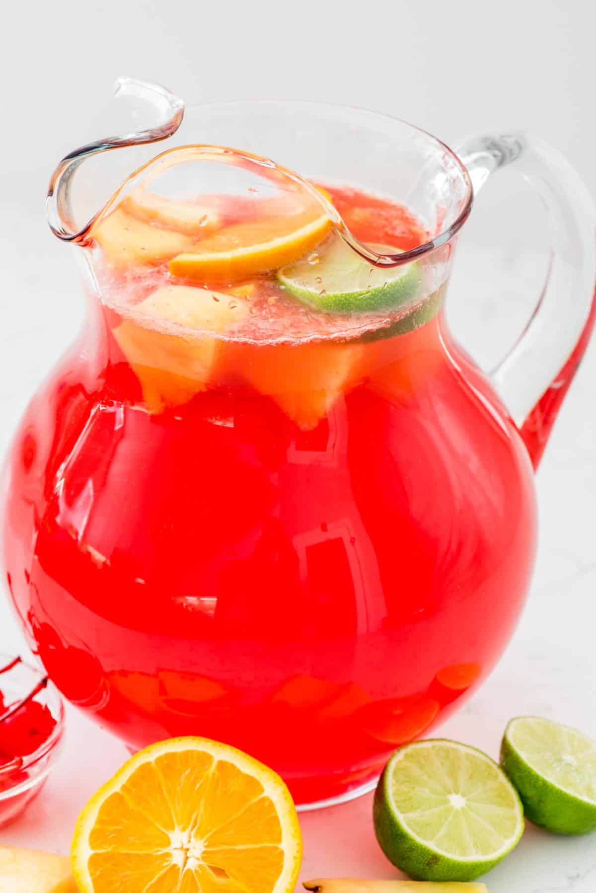 A glass pitcher filled with bright red Fruity Vodka Party Punch and slices of lemon and lime. In front, there are lemon and lime halves on a white surface.