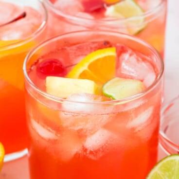Three glasses of a bright red Fruity Vodka Party Punch with ice, garnished with orange slices, lime wedges, pineapple chunks, and cherries.