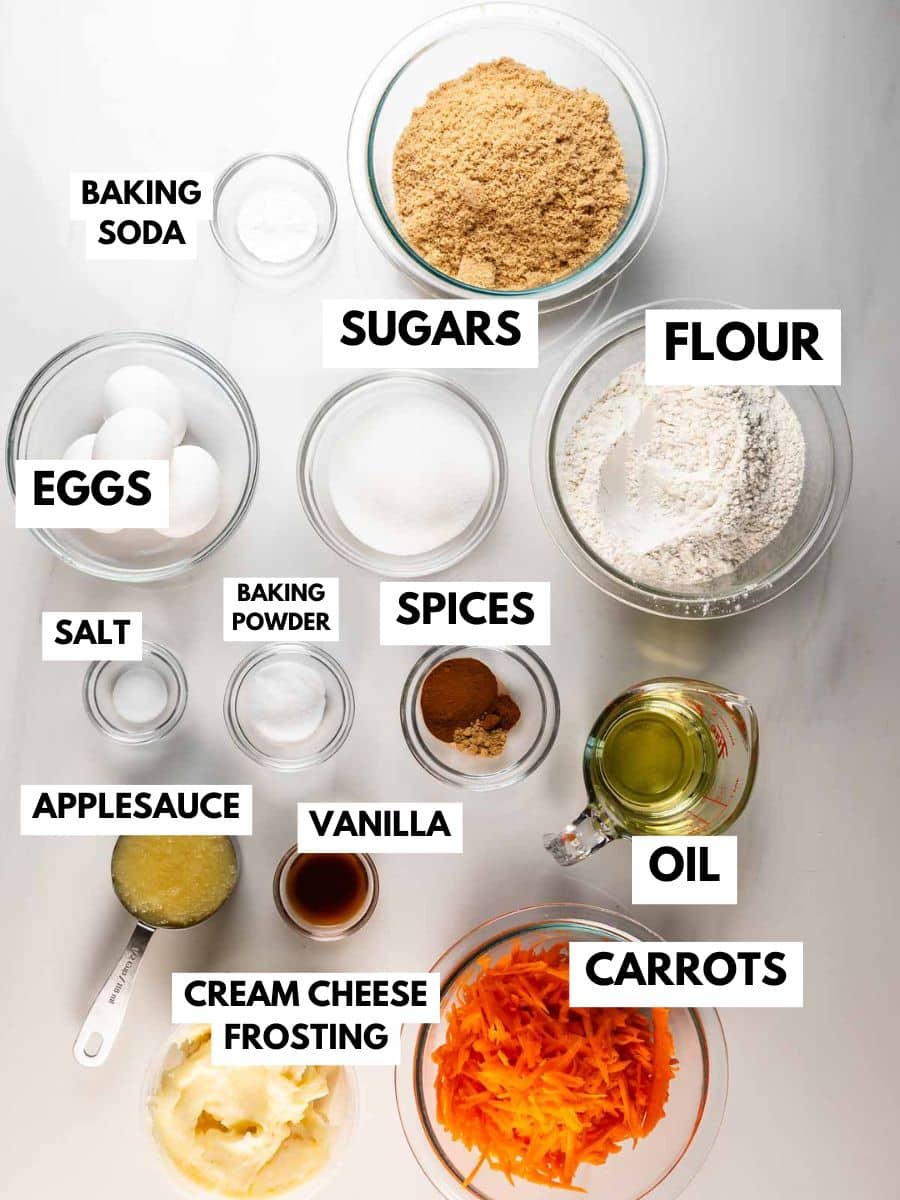 Top-down view of labeled ingredients for a Carrot Cake Loaf on a white surface, including bowls of brown and white sugar, flour, grated carrots, eggs, oil, applesauce, vanilla, spices, salt, baking soda and powder, plus cream cheese frosting.