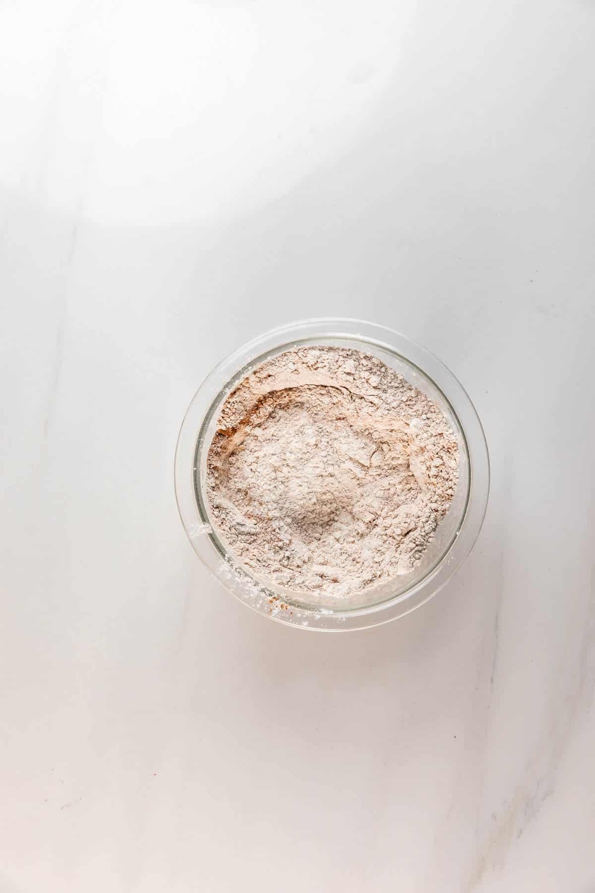 A clear glass bowl filled with a light brown flour mixture for Carrot Cake Loaf sits on a white marble countertop.