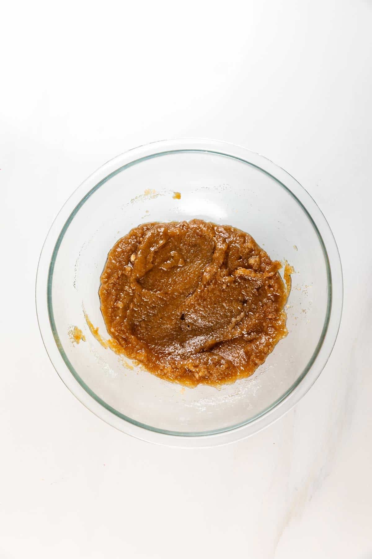 A glass bowl containing a light brown, thick cookie dough mixture with a dusting of flour on top, set on a white surface—perfect for baking a delicious Carrot Cake Loaf.