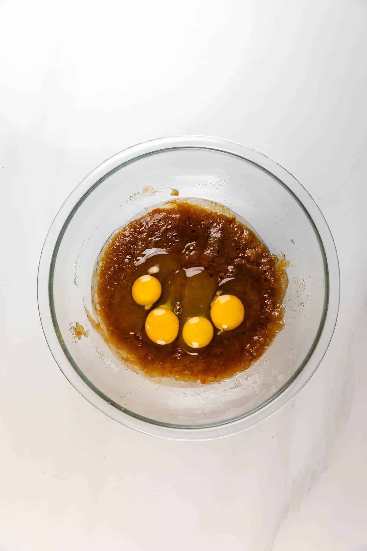 A glass mixing bowl with brown sugar mixture and four whole raw eggs on top, ready to be blended for a Carrot Cake Loaf, sits on a white countertop.
