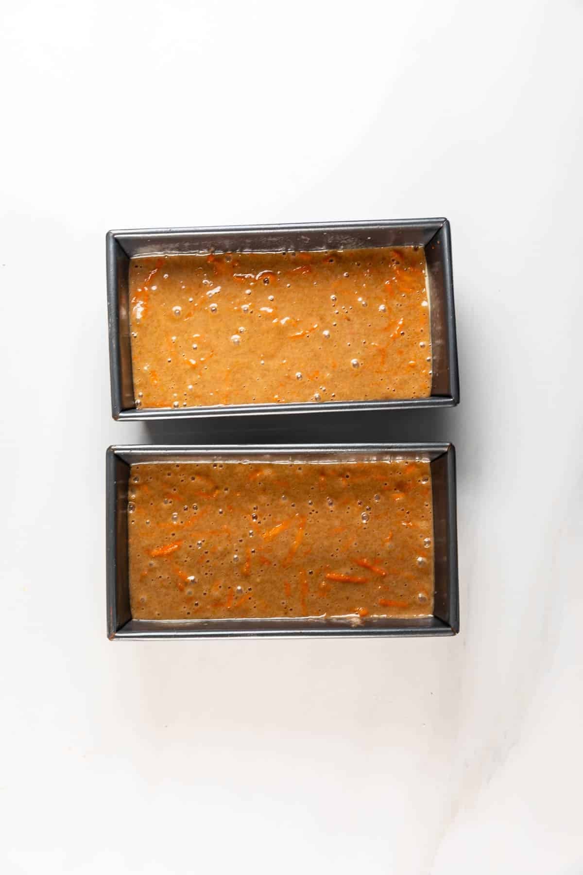 Two rectangular metal loaf pans filled with uncooked Carrot Cake Loaf batter sit on a white surface. Small bubbles and orange carrot shreds are visible in the batter, ready to be baked into moist, flavorful loaves.