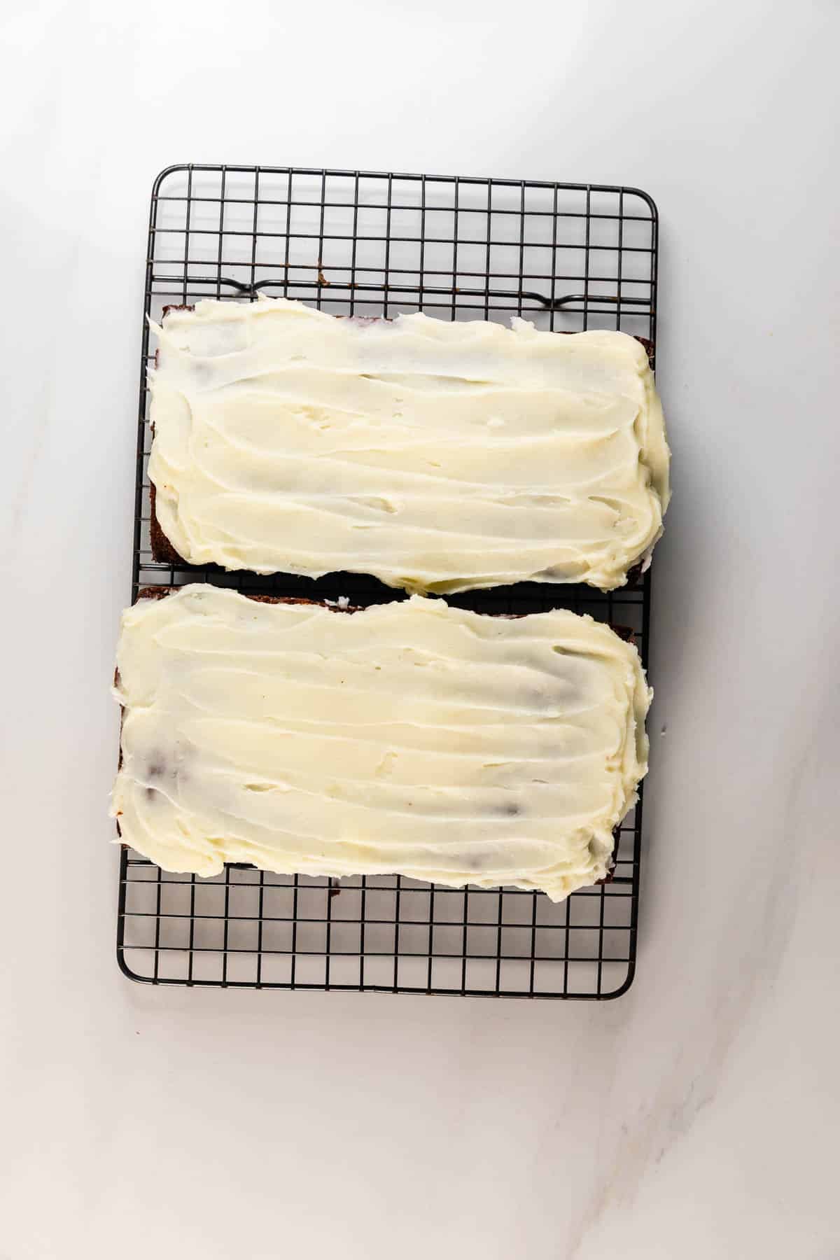 Two rectangular Carrot Cake Loaf cakes with white frosting are placed side by side on a black wire cooling rack atop a white surface.