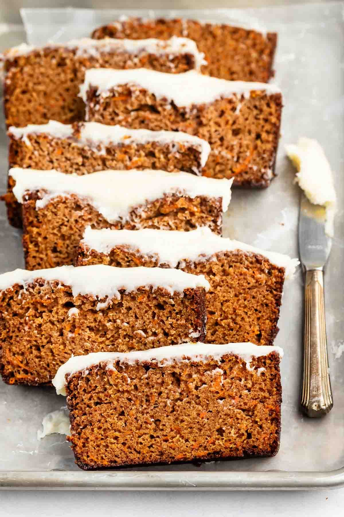 Sliced carrot cake loaf with white icing on top, arranged in a row on a baking sheet lined with parchment paper. A butter knife with icing residue rests nearby.
