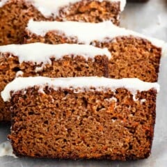 Several thick slices of moist Carrot Cake Loaf, topped with a light white icing, are arranged on a metal tray. The loaf reveals a textured, brown interior with visible orange specks of carrot throughout.