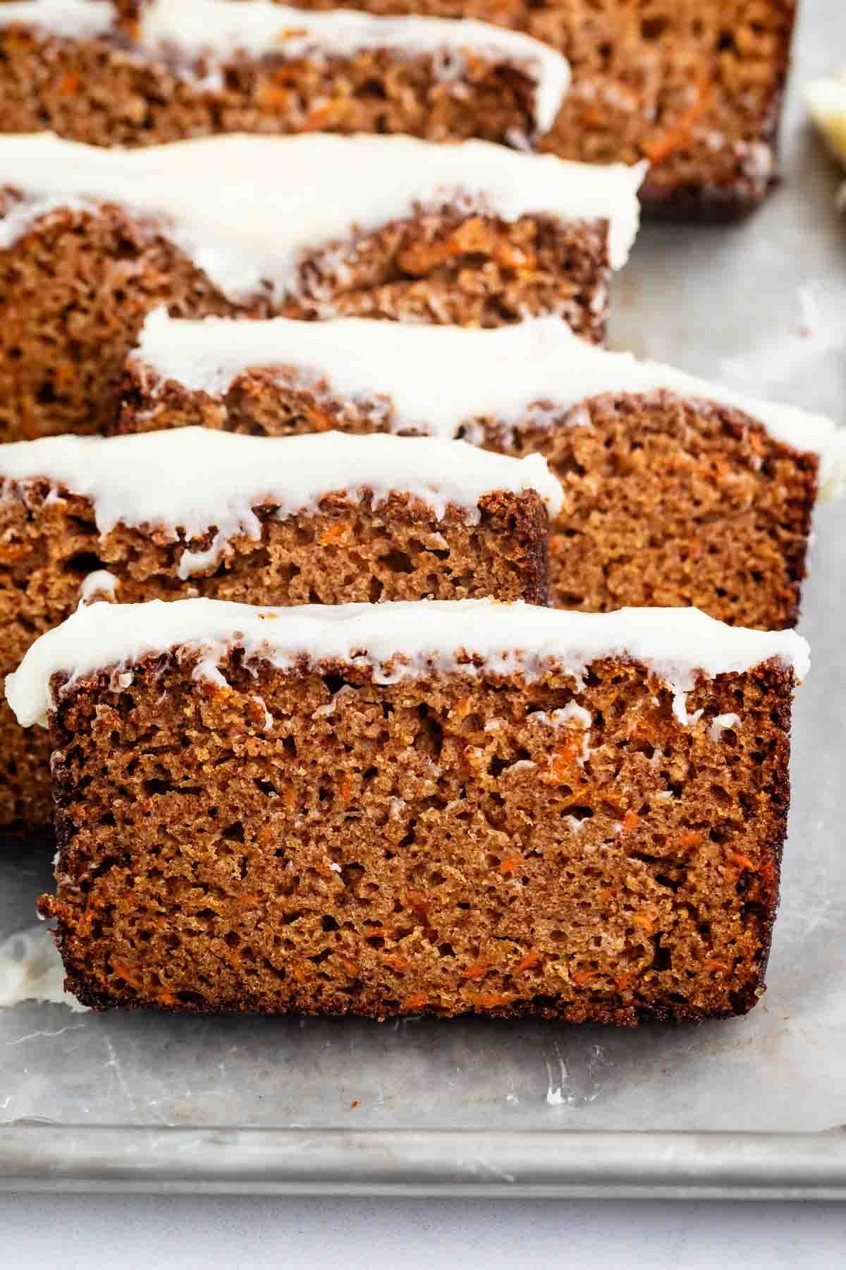 Several thick slices of moist Carrot Cake Loaf, topped with a light white icing, are arranged on a metal tray. The loaf reveals a textured, brown interior with visible orange specks of carrot throughout.