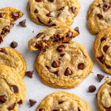 A close-up of several chocolate chip cookies on a light surface, with some cookies broken in half to reveal the gooey centers—a perfect inspiration for your next Chocolate Chip Cookie Recipe. Scattered chocolate chips complete the delicious scene.