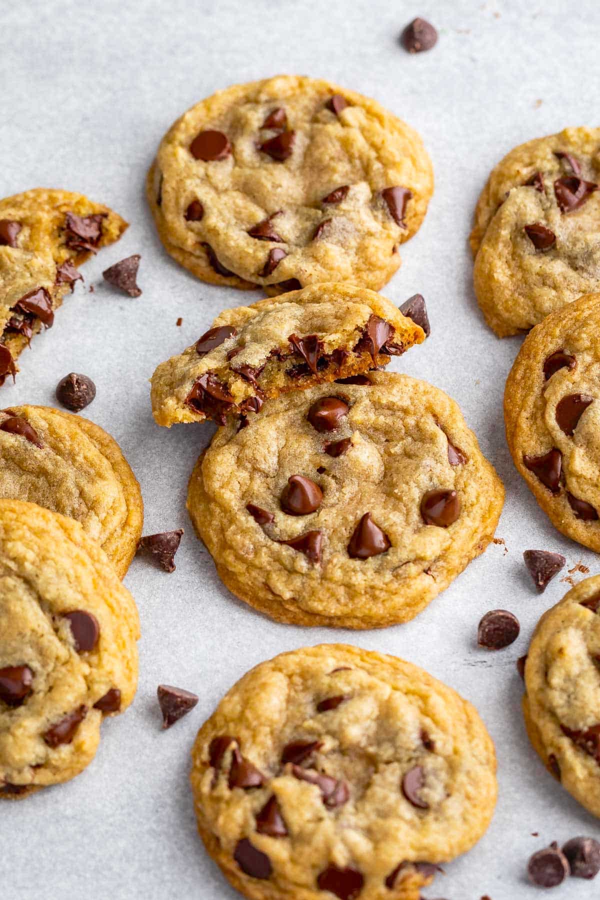 A close-up of several chocolate chip cookies on a light surface, with some cookies broken in half to reveal the gooey centers—a perfect inspiration for your next Chocolate Chip Cookie Recipe. Scattered chocolate chips complete the delicious scene.