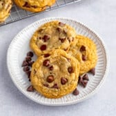 Three chocolate chip cookies sit on a white plate with a few chocolate chips scattered around them—a tempting preview of this delicious Chocolate Chip Cookie Recipe. In the background, more cookies cool on a rack.
