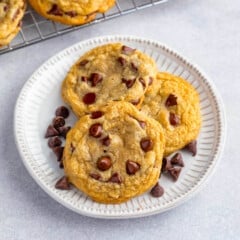 Three chocolate chip cookies sit on a white plate with a few chocolate chips scattered around them—a tempting preview of this delicious Chocolate Chip Cookie Recipe. In the background, more cookies cool on a rack.