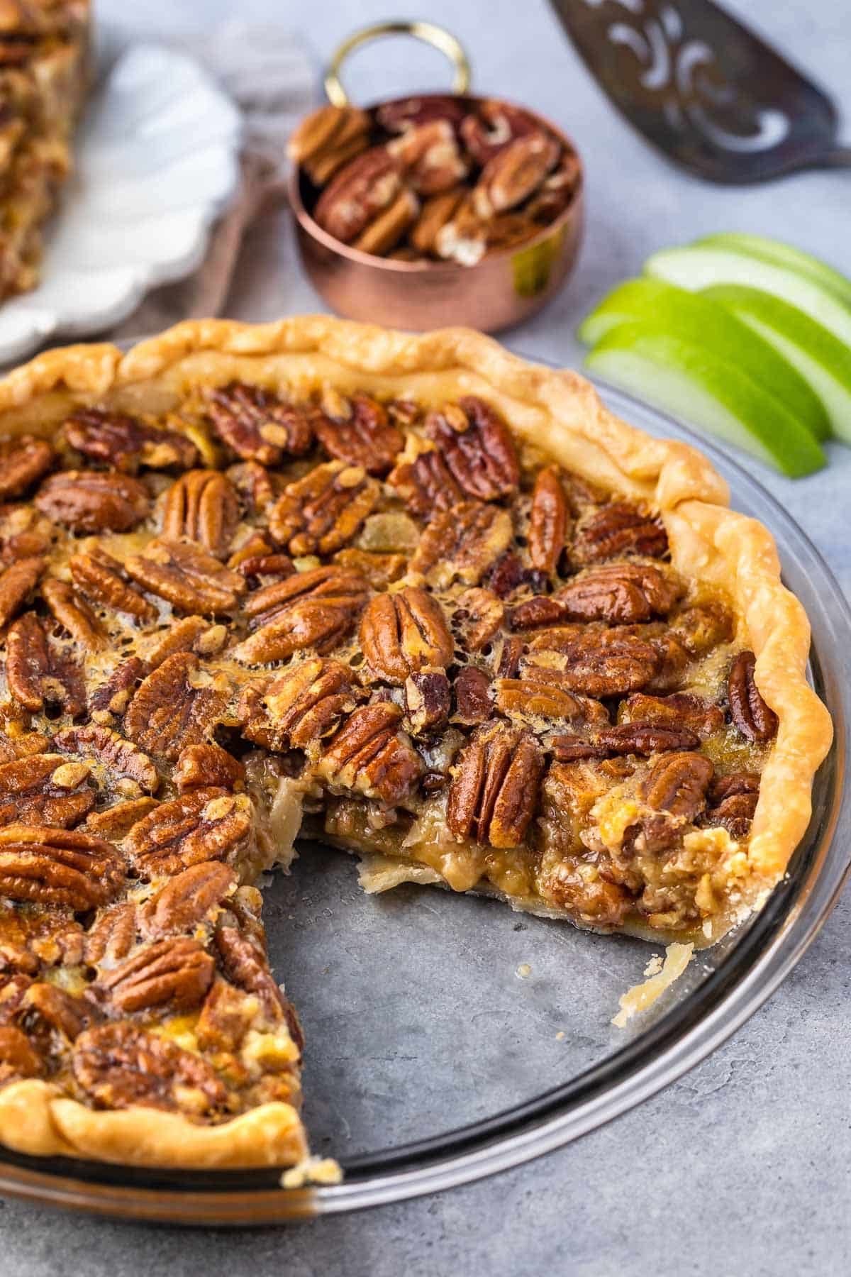 pie with slice missing, sliced apples in back and a cup of pecans