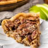 one slice of pecan pie on a white plate with sliced apples in the back