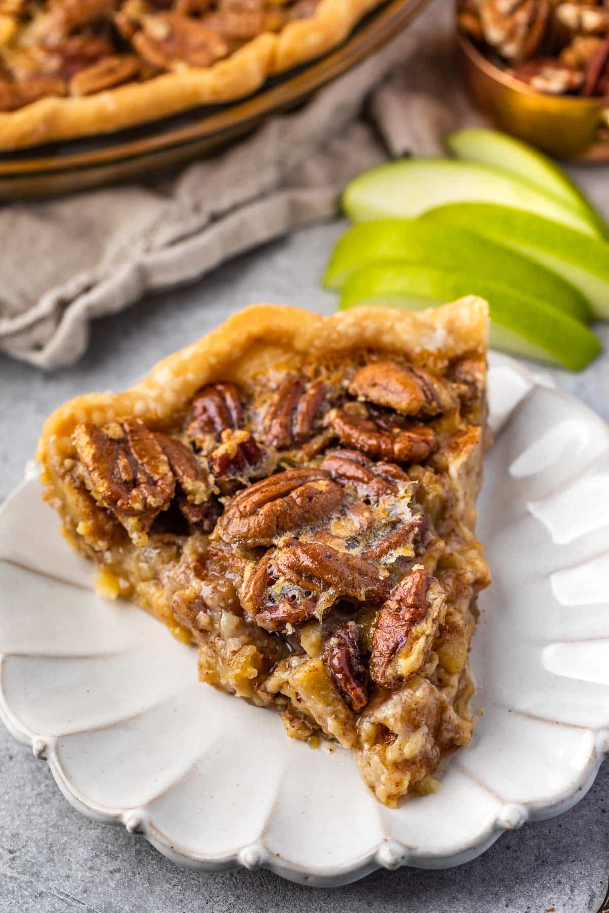 one slice of pecan pie on a white plate with sliced apples in the back 
