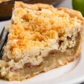 A close-up of a slice of Apple Crumb Pie on a white plate with a fork beside it. The pie has a golden, crumbly topping and a visible apple filling.