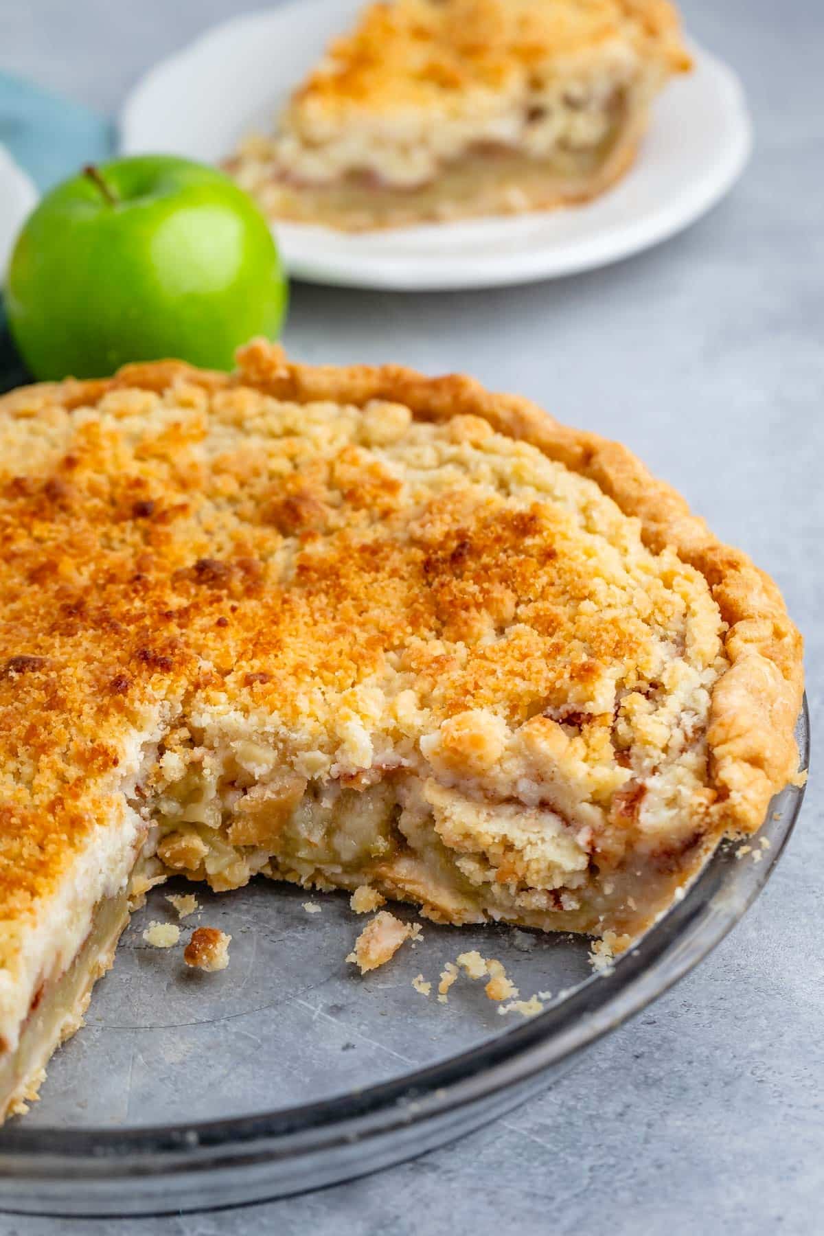 A delicious Apple Crumb Pie with a golden crumb topping sits in a glass pie dish with a slice missing; a green apple and a plated slice of pie are in the background on a gray surface.