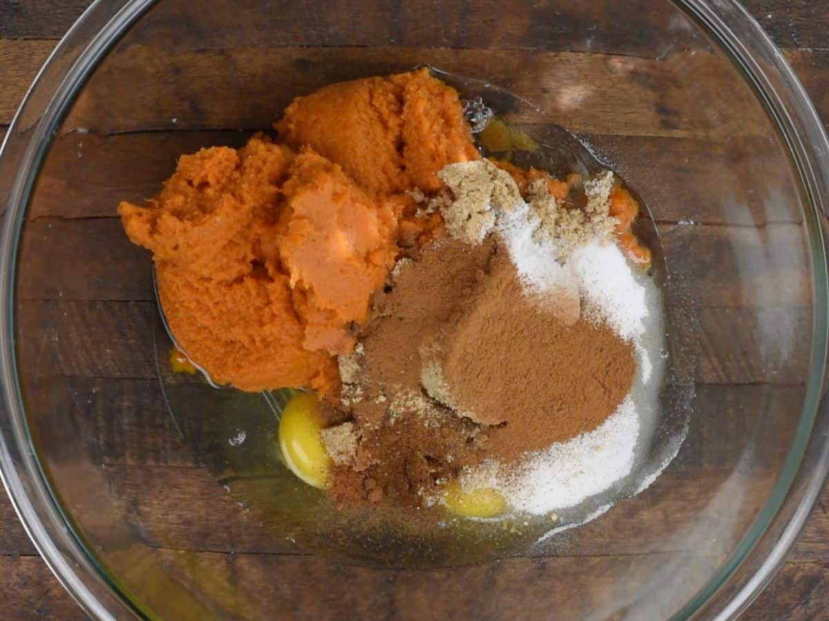 pumpkin, sugar, eggs, spices in large glass bowl on wood table.