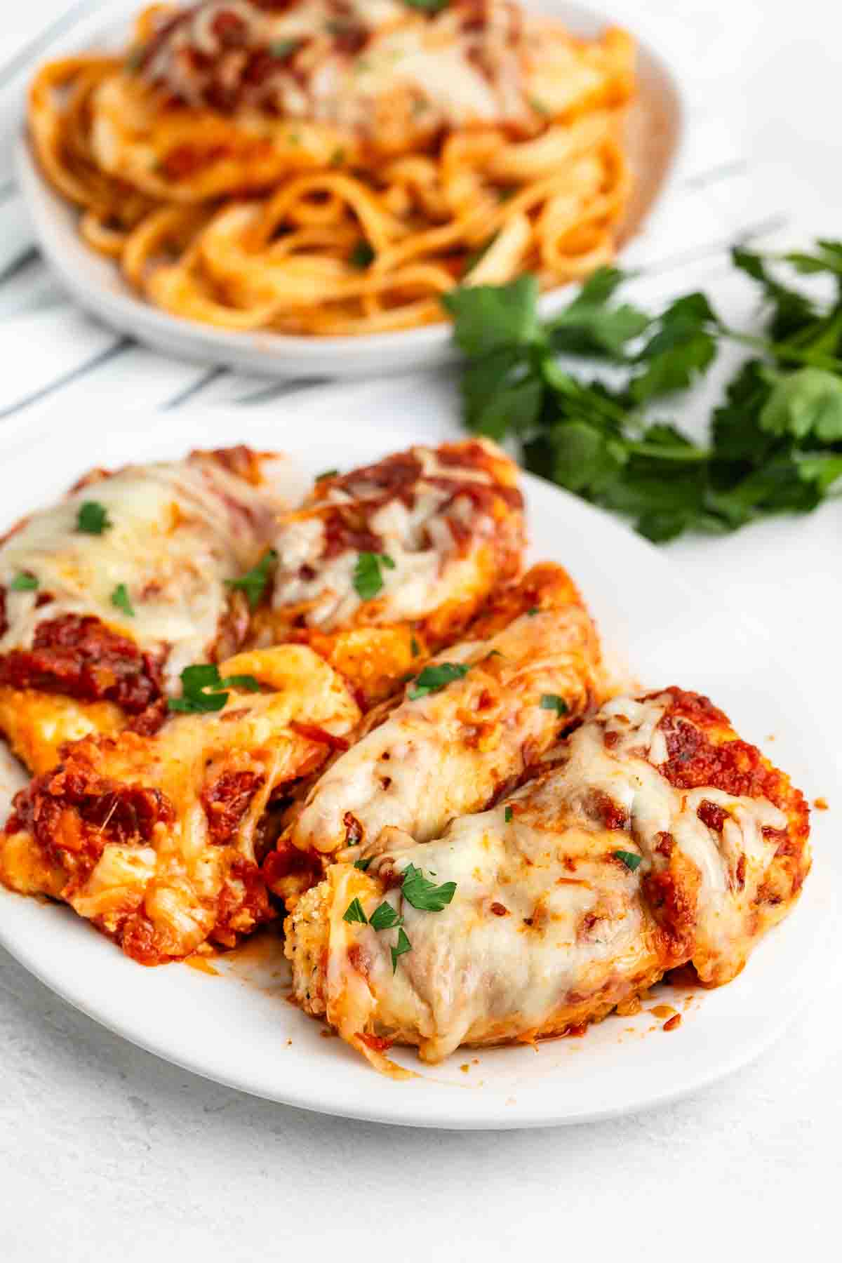 A white plate features an easy chicken Parmesan recipe: breaded chicken breasts topped with melted cheese and marinara sauce, garnished with parsley. In the background, a plate of pasta and a bunch of fresh parsley are visible.