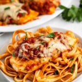 A plate of fettuccine pasta topped with a baked chicken breast, marinara sauce, and melted cheese, garnished with chopped parsley—an easy chicken Parmesan recipe. More chicken is served on a platter in the background.