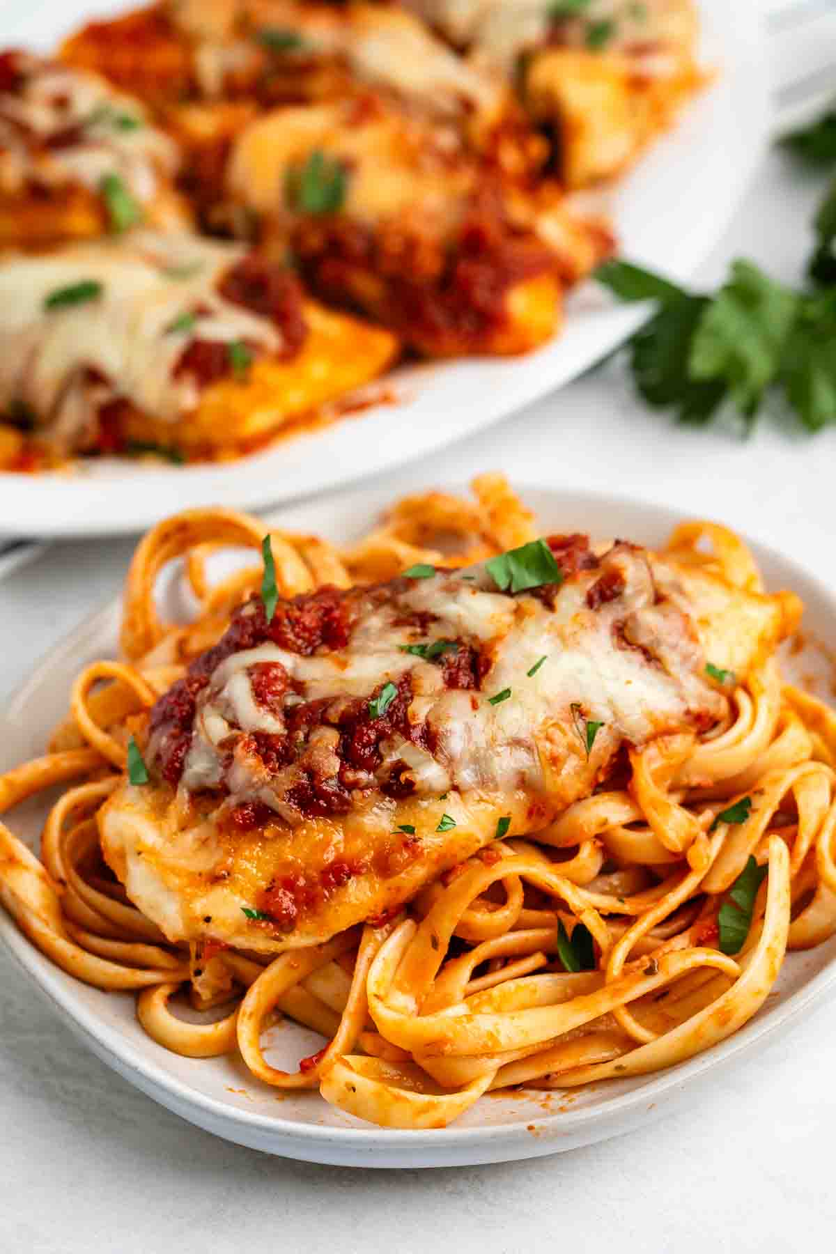 A plate of fettuccine pasta topped with a baked chicken breast, marinara sauce, and melted cheese, garnished with chopped parsley—an easy chicken Parmesan recipe. More chicken is served on a platter in the background.