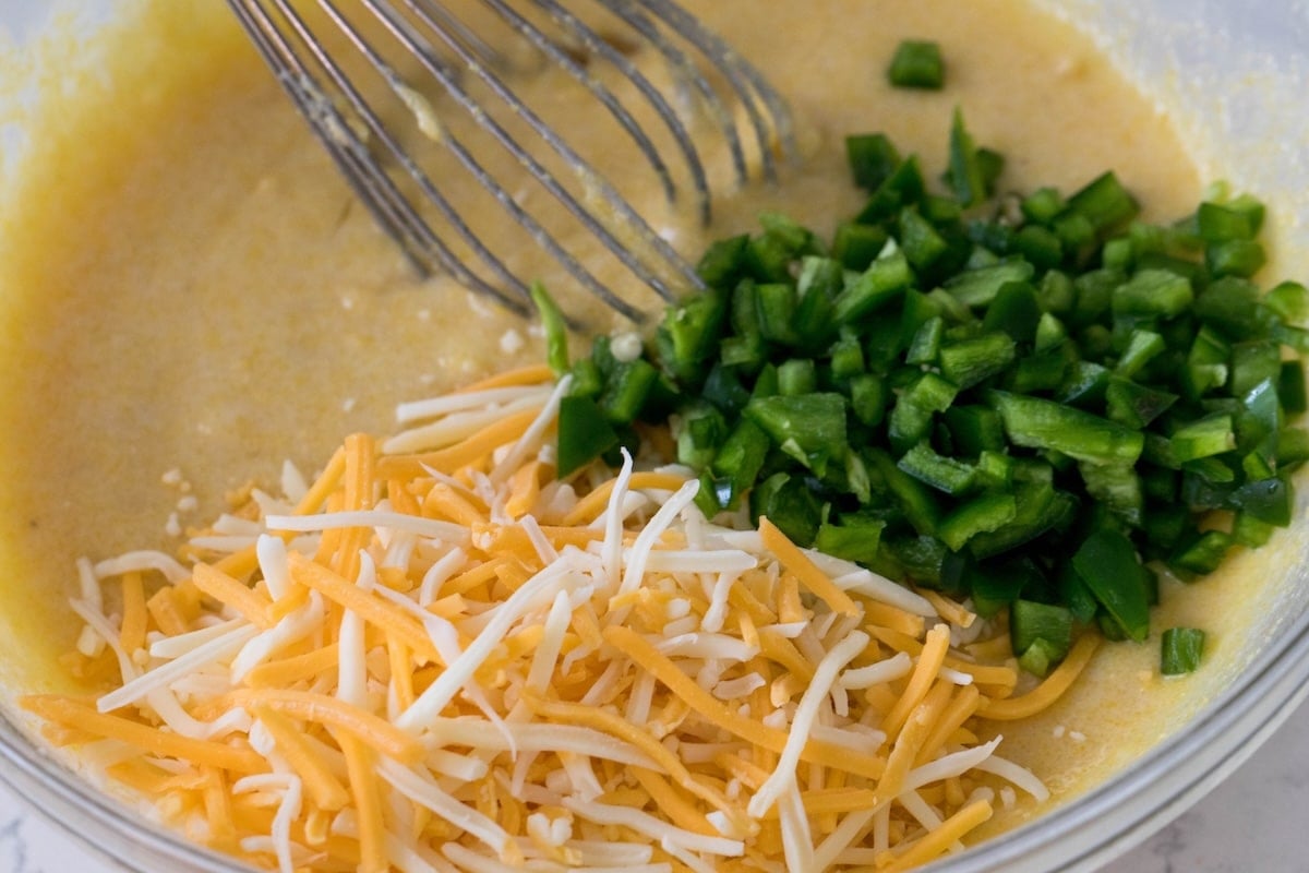 A mixing bowl brims with Mexican cornbread batter, featuring shredded cheddar, mozzarella cheese, and chopped green bell peppers. A metal whisk rests in the mixture, ready to bring these vibrant ingredients together.
