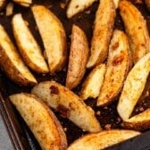 Seasoned oven roasted potatoes cut into wedges and baked on a dark tray, golden brown with crispy edges and visible spices, arranged in a single layer.