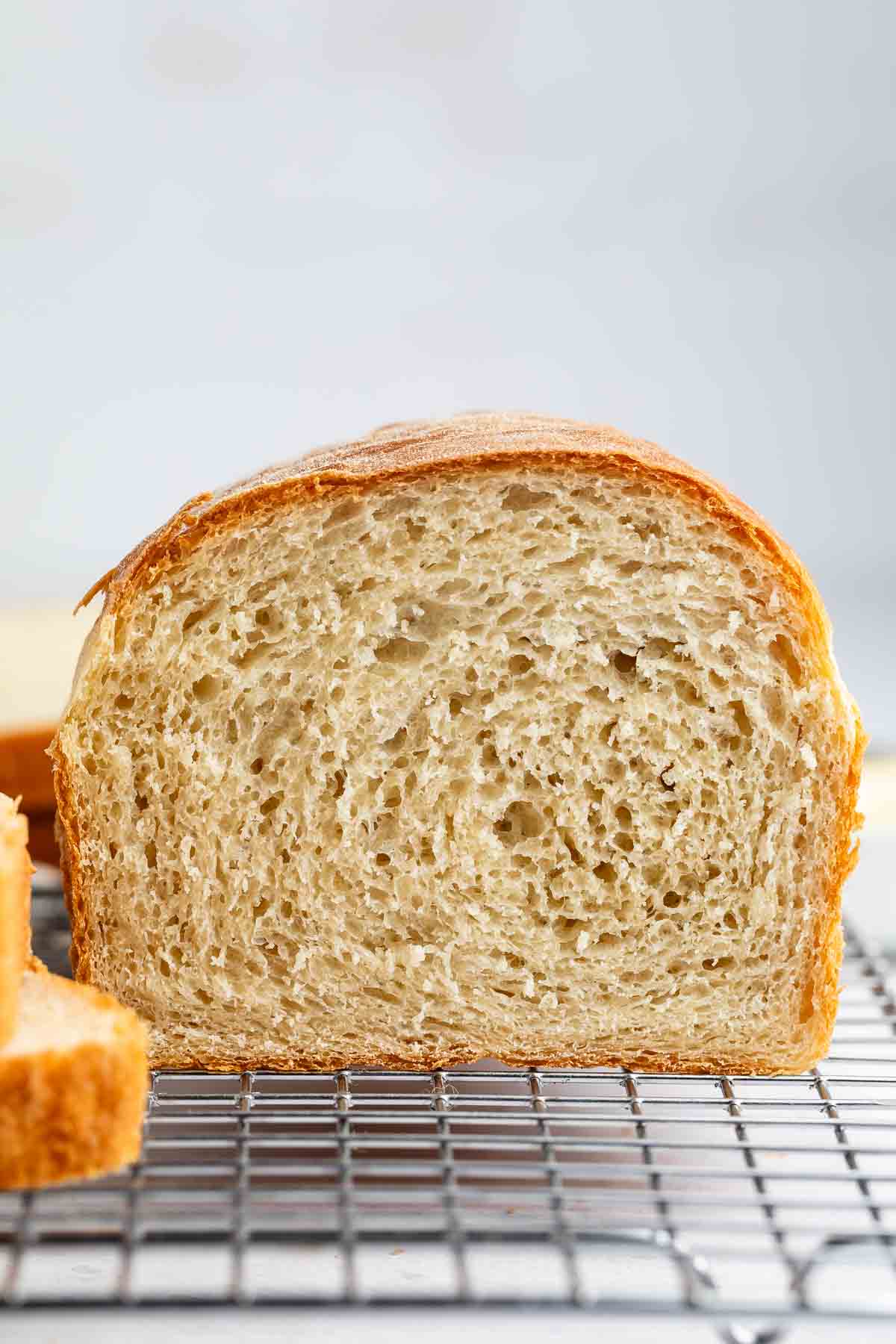 A loaf of homemade white bread with a golden-brown crust and soft, airy interior sits on a metal cooling rack, with a slice cut from one end.