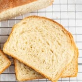 Two slices of homemade white bread rest on a cooling rack, with part of the loaf visible above them. The bread features a golden-brown crust and a soft, airy crumb.