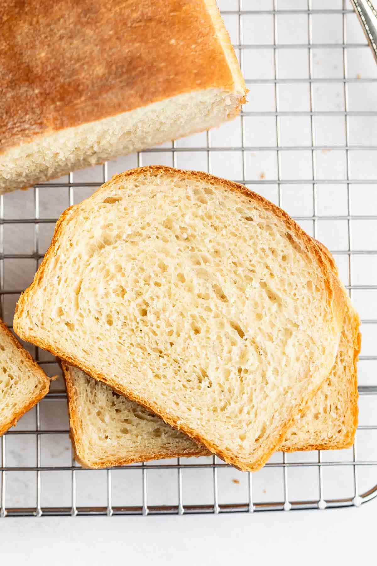 Two slices of homemade white bread rest on a cooling rack, with part of the loaf visible above them. The bread features a golden-brown crust and a soft, airy crumb.