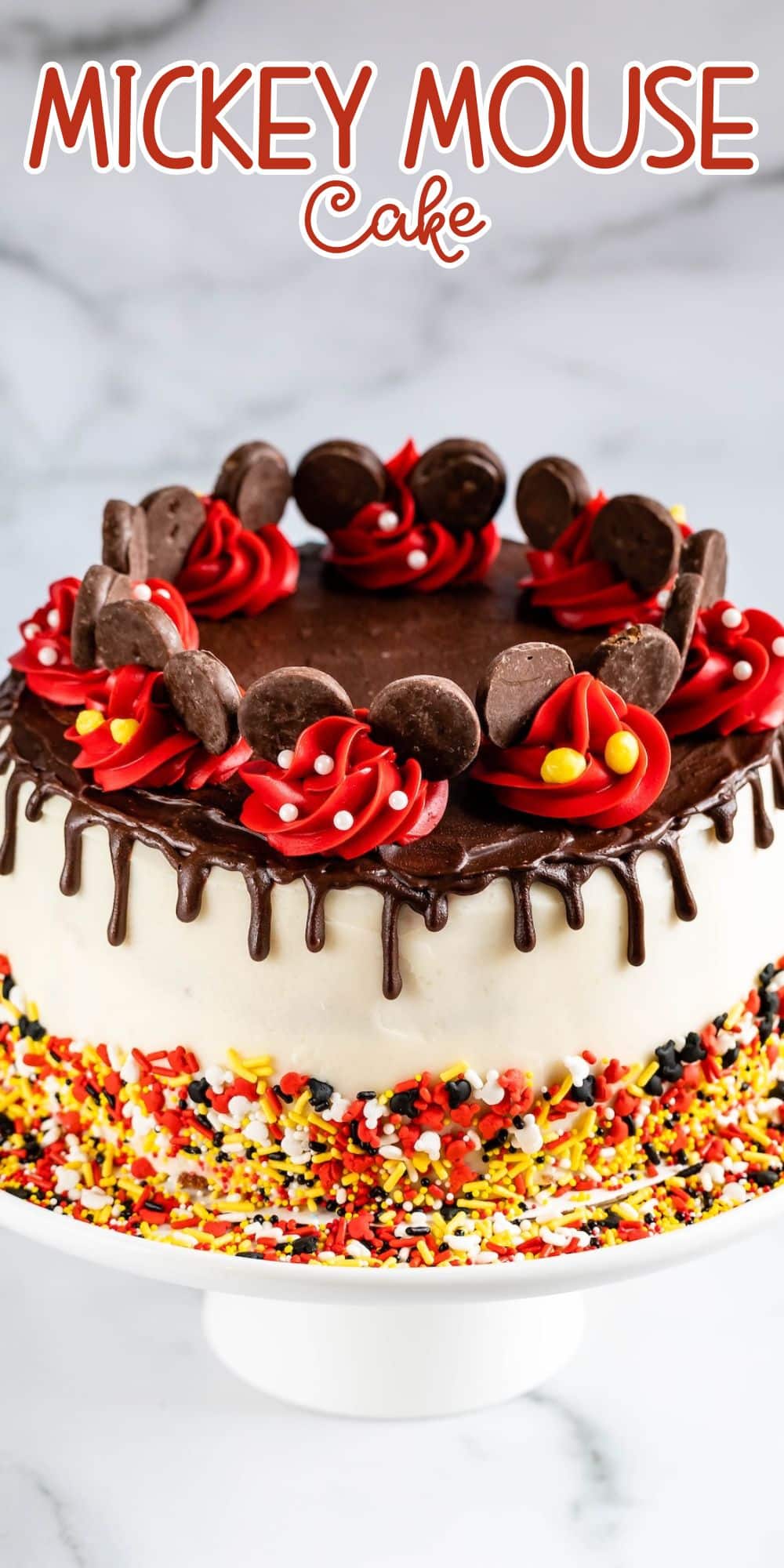 A festive Mickey Mouse cake decorated with chocolate glaze, red and yellow frosting, chocolate cookie ears, and colorful sprinkles, displayed on a white cake stand. Text above reads Mickey Mouse Cake.