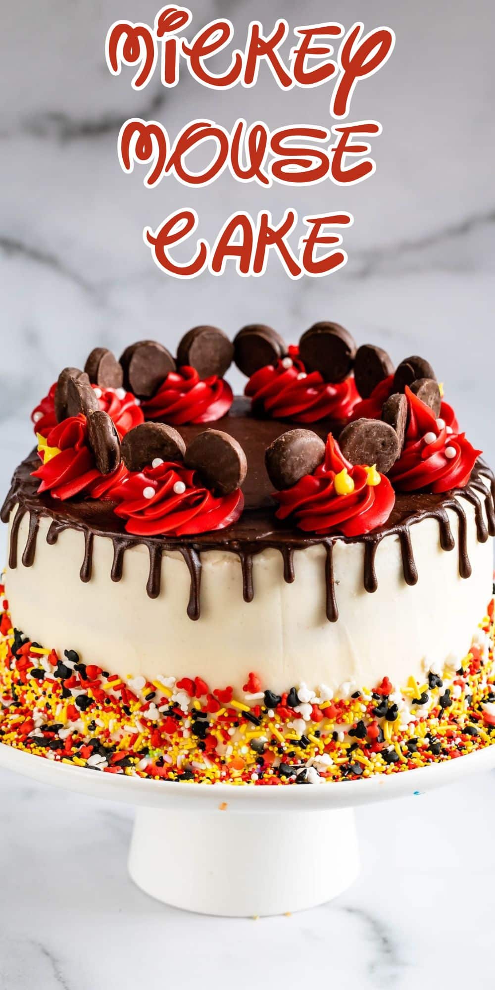 A round Mickey Mouse cake decorated with white frosting, chocolate drip, red and yellow icing, chocolate disks, and colorful sprinkles sits on a white cake stand. The text Mickey Mouse Cake appears above the cake.