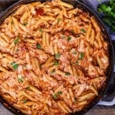 A skillet filled with chicken penne pasta, shredded chicken, and marinara sauce, garnished with fresh parsley, sits on a wooden surface next to a bunch of parsley and a wedge of cheese.